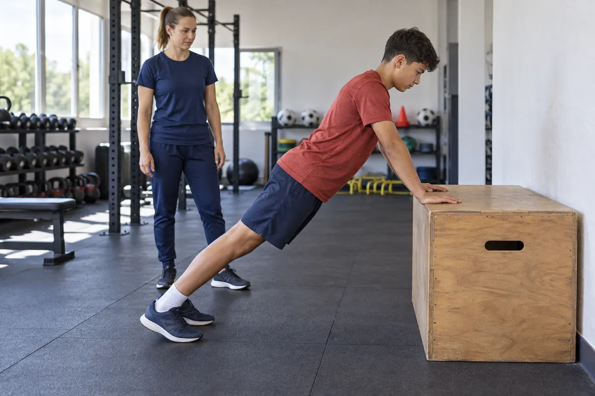 Ein Jugendlicher stützt die Hände auf eine stabile hohe Trainingsbox; eine Trainerin beobachtet die Ausgangsposition